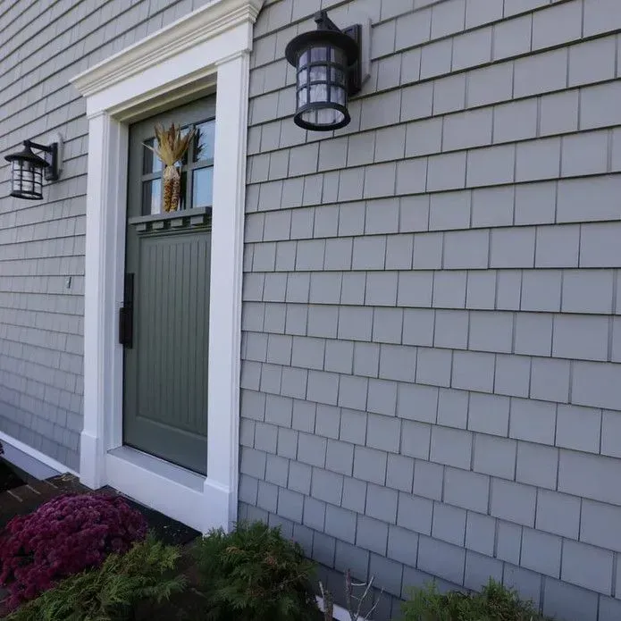 A sage green front door with a glass pane, set in gray shingle siding with white trim and black outdoor lanterns.