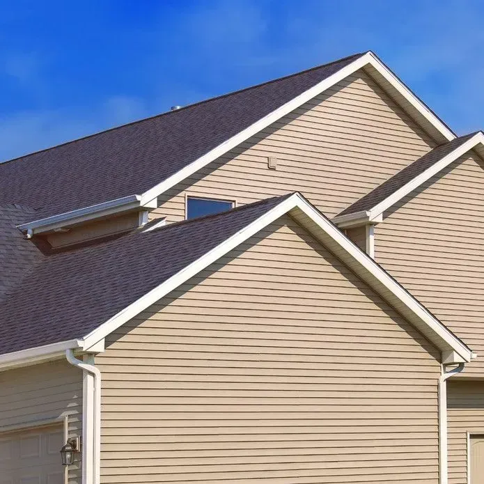 Tan vinyl siding covers the gables and walls of a multi-level house with a dark shingled roof under a clear blue sky.