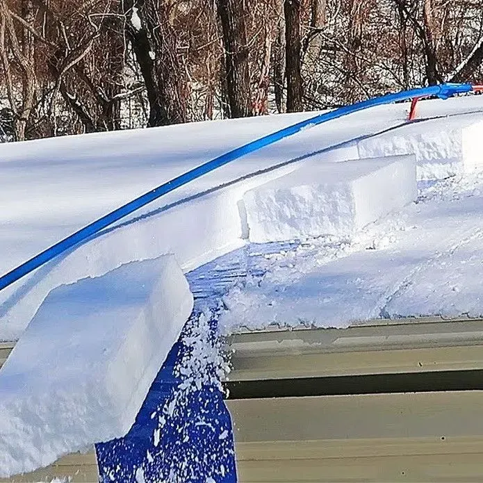 A blue roof rake tool is used to clear blocks of snow from a metal roof covered in deep snow near bare winter trees.