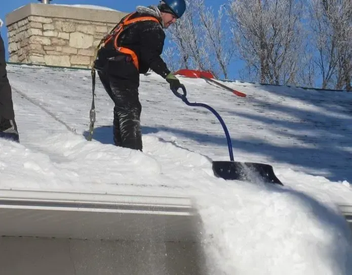 A person in safety gear uses a blue-handled tool to clear deep snow from a residential roof on a sunny day.