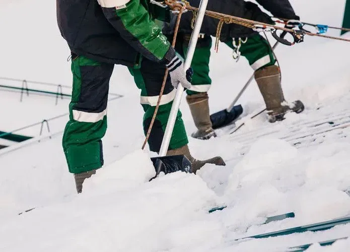 Two workers in green and black uniforms use long-handled shovels to clear snow from a sloped roof while secured by ropes.
