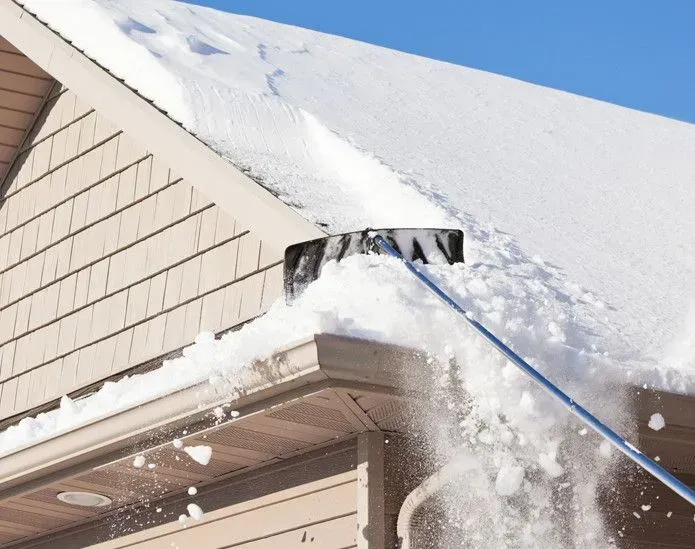 A snow rake clears a thick layer of snow off the roof of a beige-sided house against a bright blue sky.
