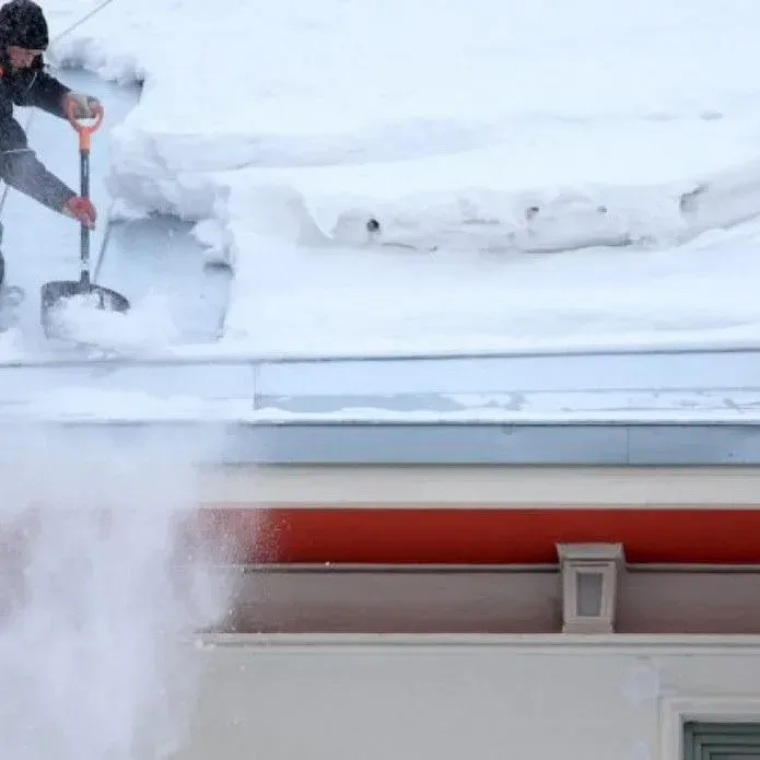 A person in dark clothing clears snow from a metal roof using a shovel, sending snow falling over the edge.