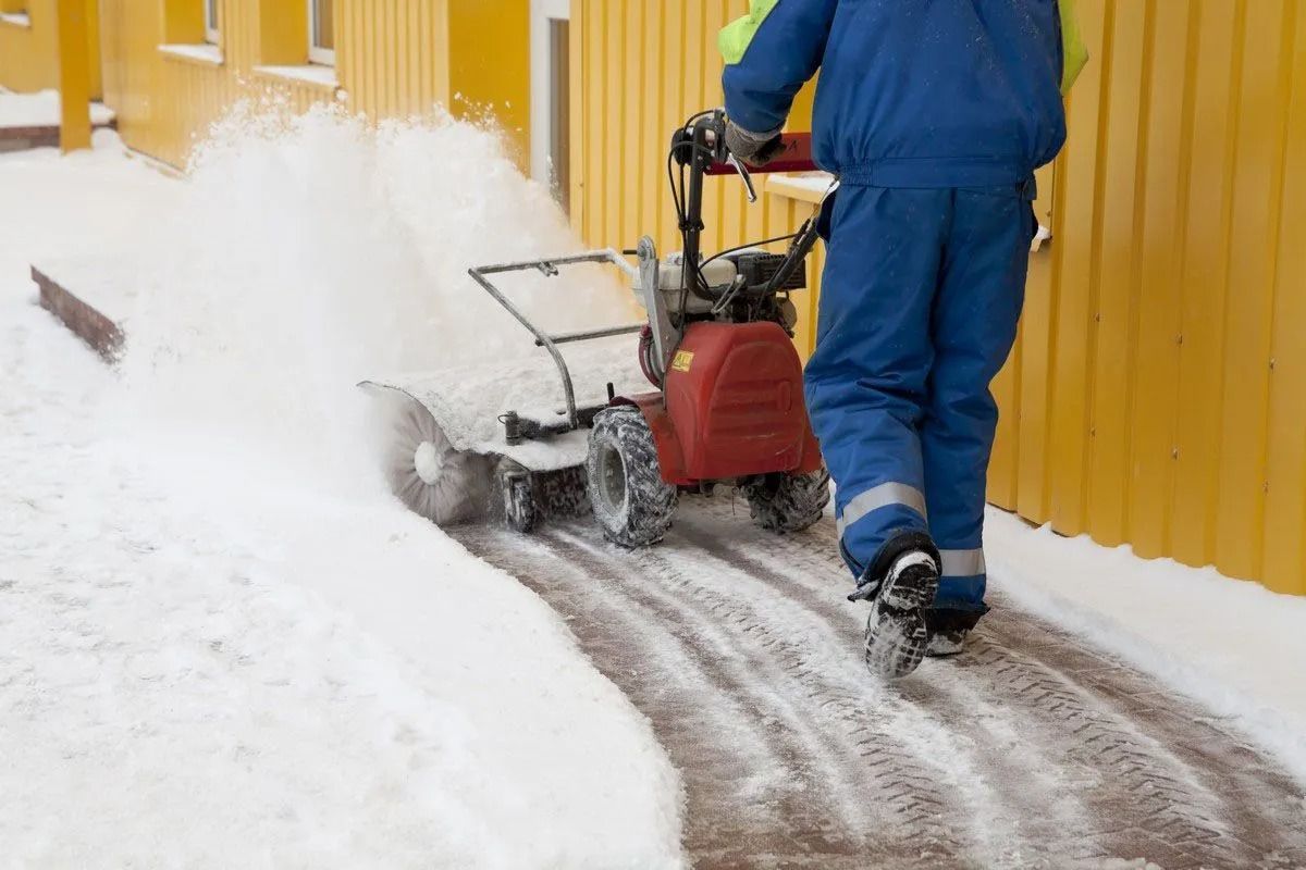 A person in blue coveralls uses a red motorized snow sweeper to clear a path along a bright yellow building.