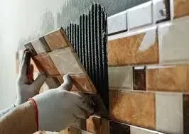 A person wearing gloves presses a rectangular, multi-toned brown tile onto a wall covered in adhesive ridges.