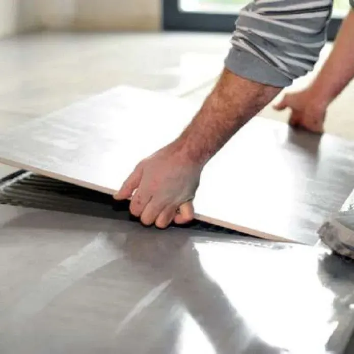 A person’s hands placing a large square floor tile onto a surface prepared with adhesive mortar.