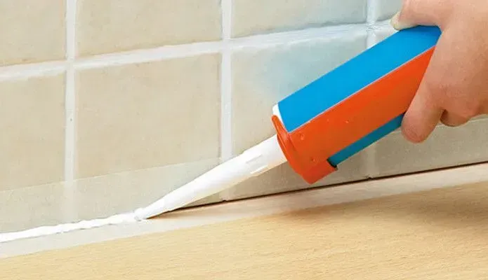 A hand uses a caulk gun to apply a bead of white sealant along the joint between tiled wall and a wooden surface.