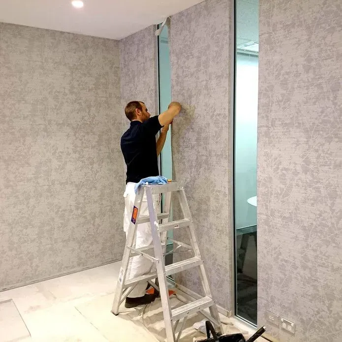 A worker standing on a ladder applies gray textured wallpaper to a wall next to a glass partition.