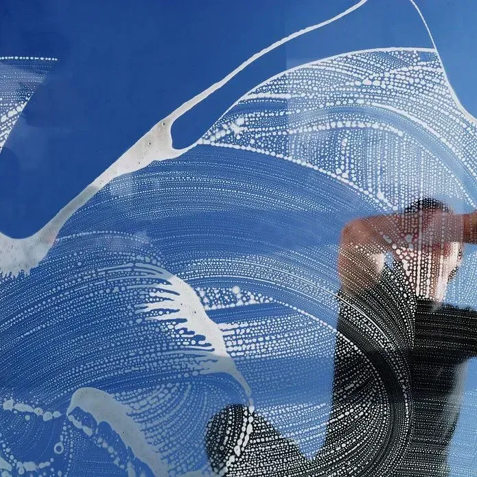 A person in a black shirt cleans a window covered in white soap suds, revealing a clear blue sky through the glass.