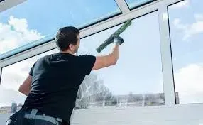 A person in a black t-shirt uses a squeegee to clean a large glass window outdoors under a blue sky.