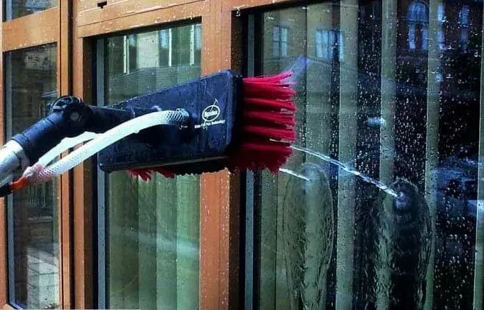 A water-fed cleaning brush with red bristles sprays water onto a glass window pane set in a wooden frame.