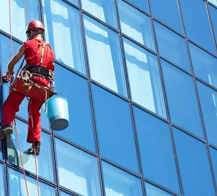 A person in red coveralls and a helmet rappels down a modern glass skyscraper, carrying a blue bucket while cleaning windows.