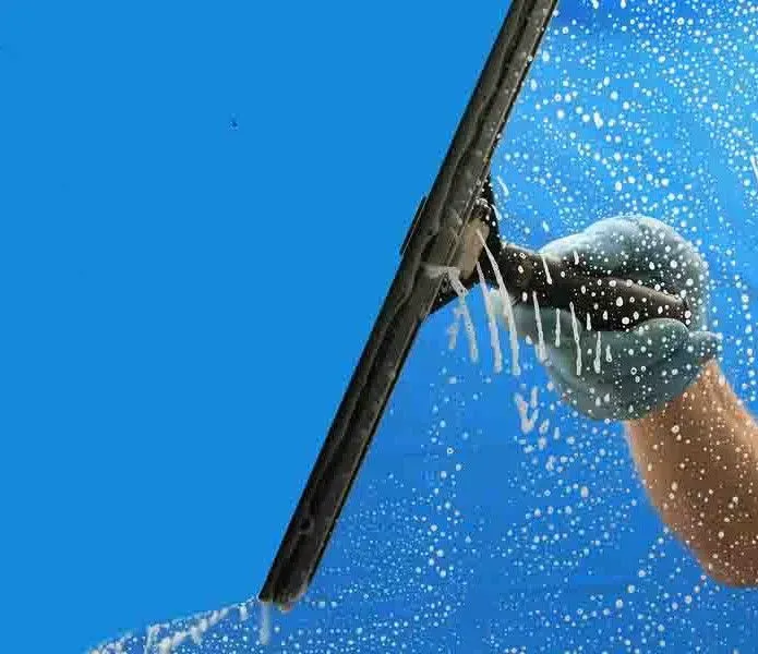 A gloved hand uses a squeegee to wipe soapy water off a glass surface against a clear blue sky.