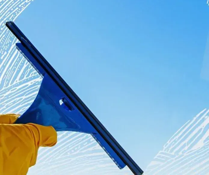 A yellow-gloved hand uses a blue squeegee to wipe cleaning suds off a glass window against a clear blue sky.