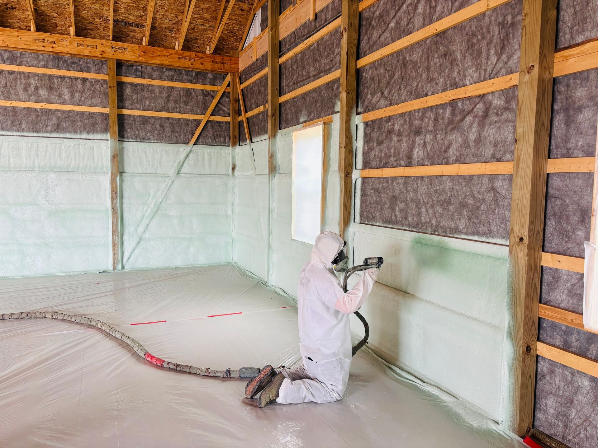 Person in protective suit spraying insulation into a building's walls and floor. Interior view with wood framing.