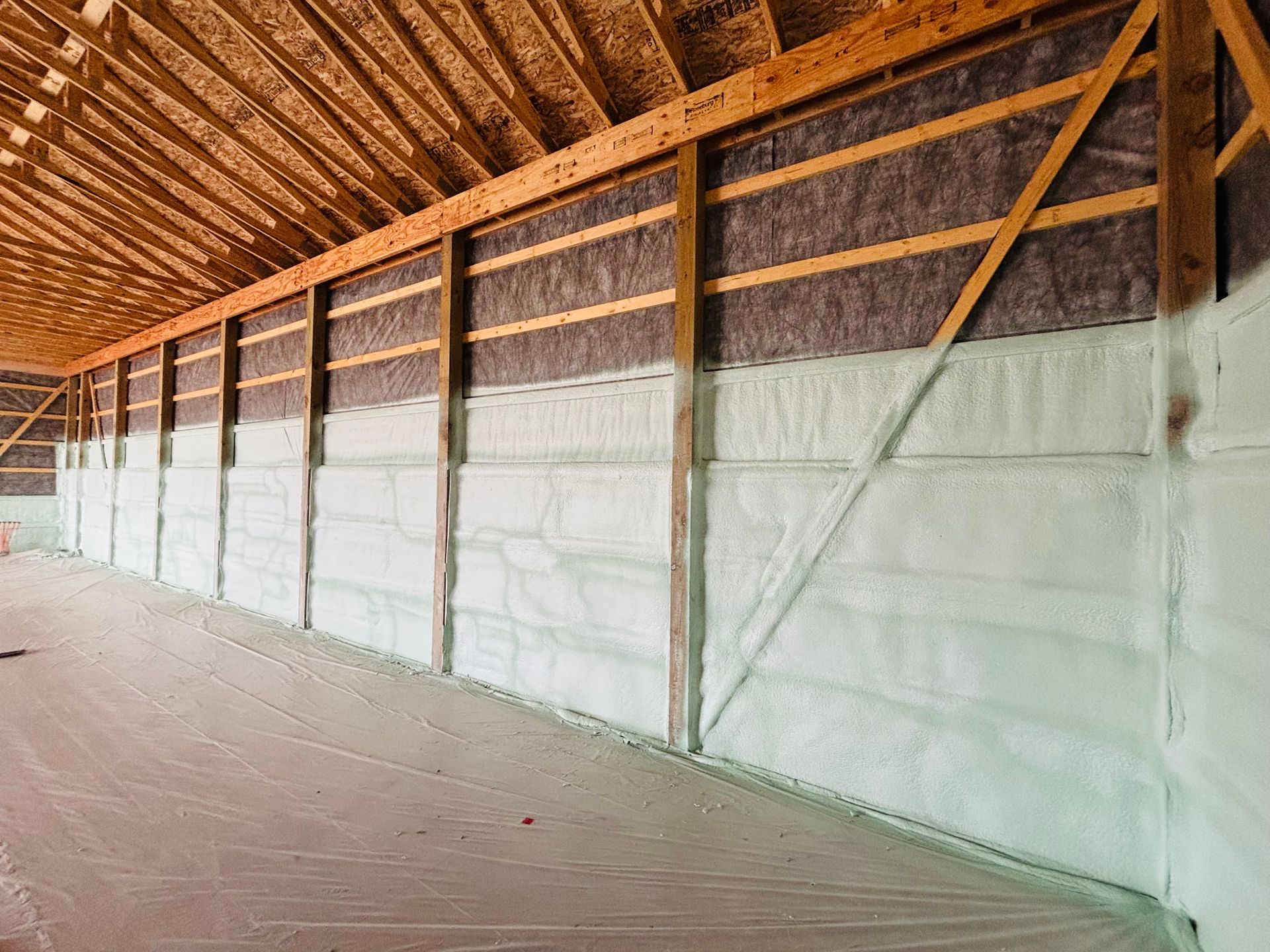 Interior of a building under construction, walls insulated with white spray foam and dark batting; wooden beams visible.