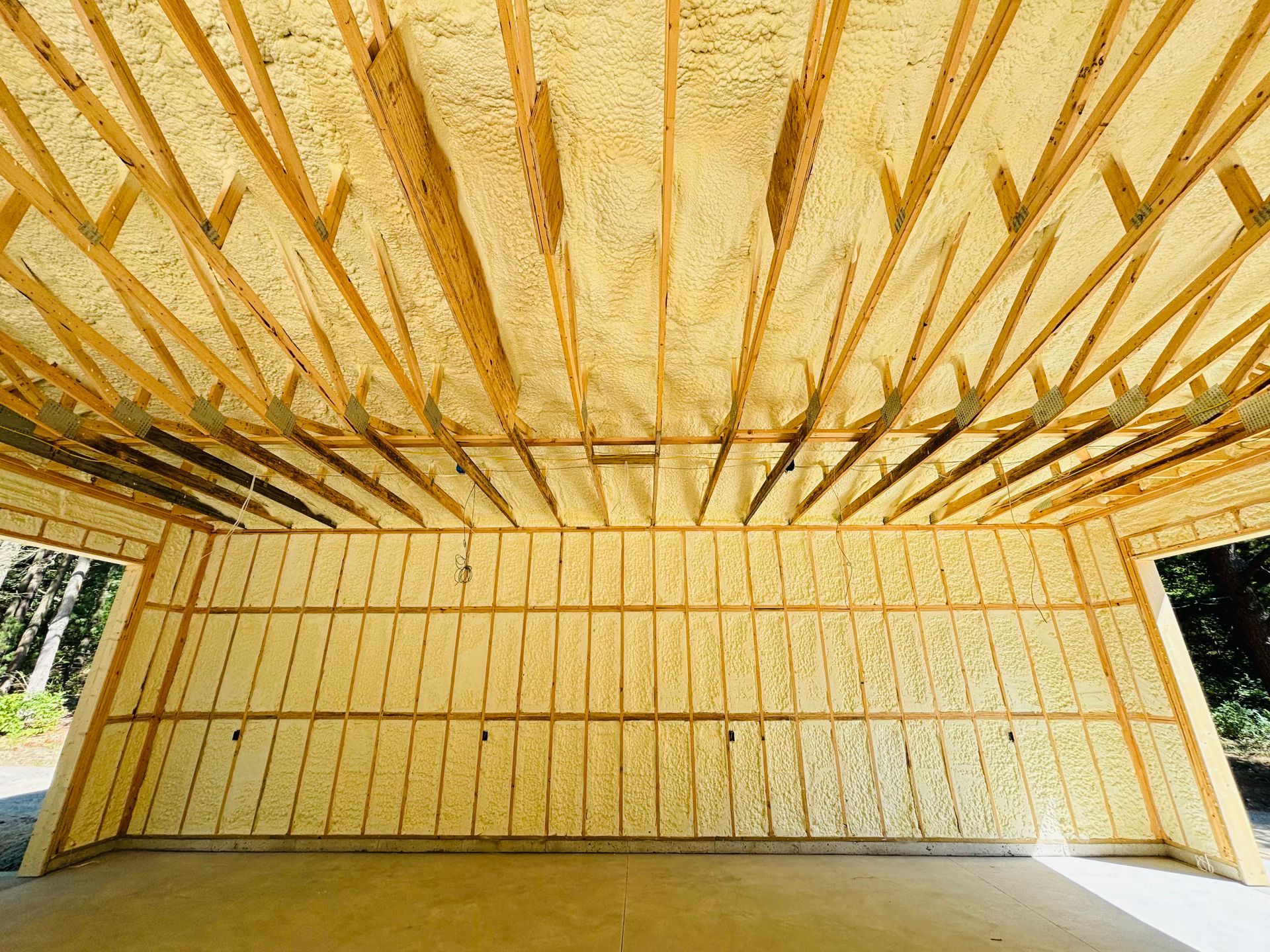 Garage interior under construction, filled with yellow spray foam insulation between wooden beams and rafters.