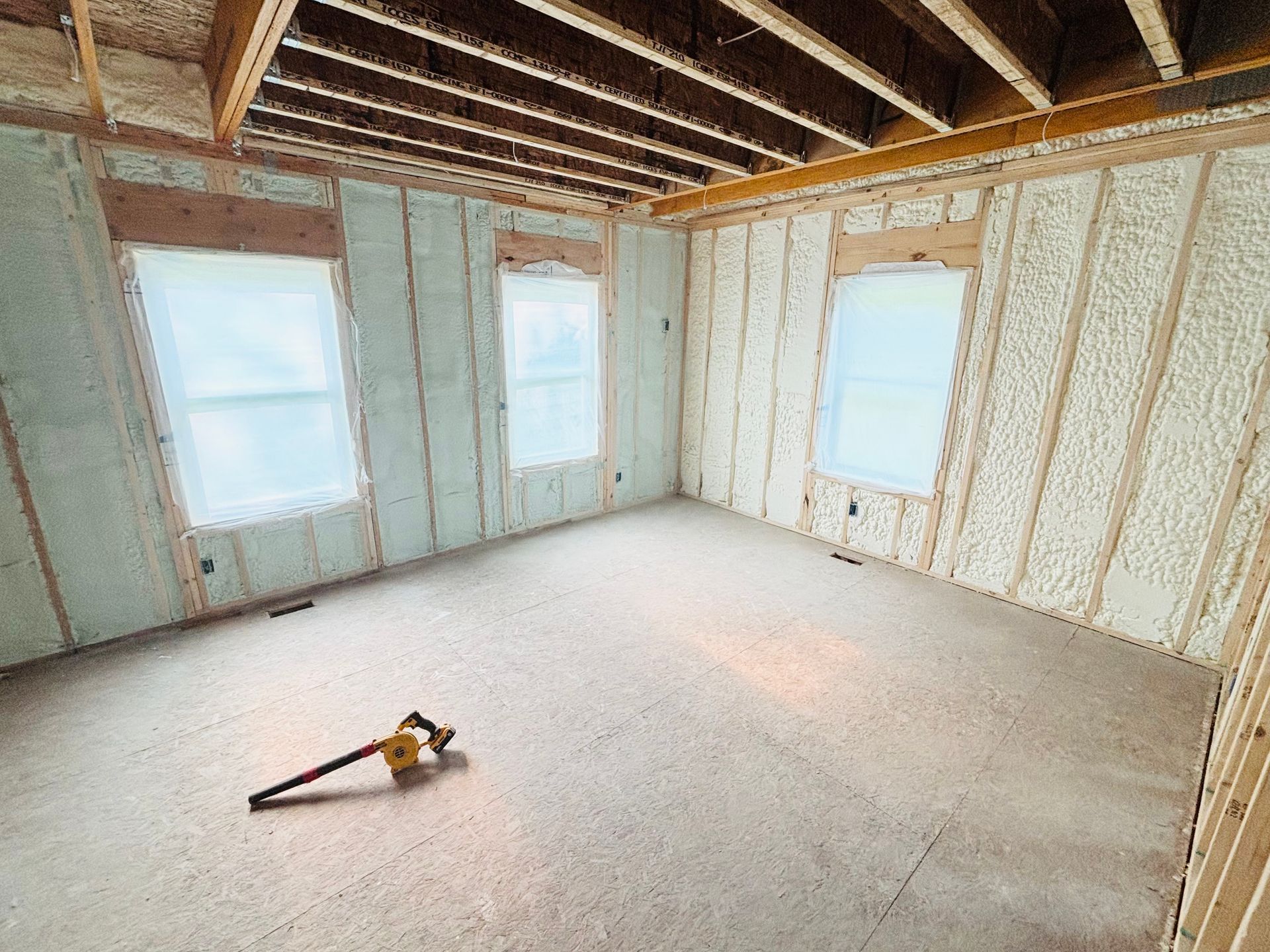 Interior room under construction, with spray foam insulation visible on walls; windows present.