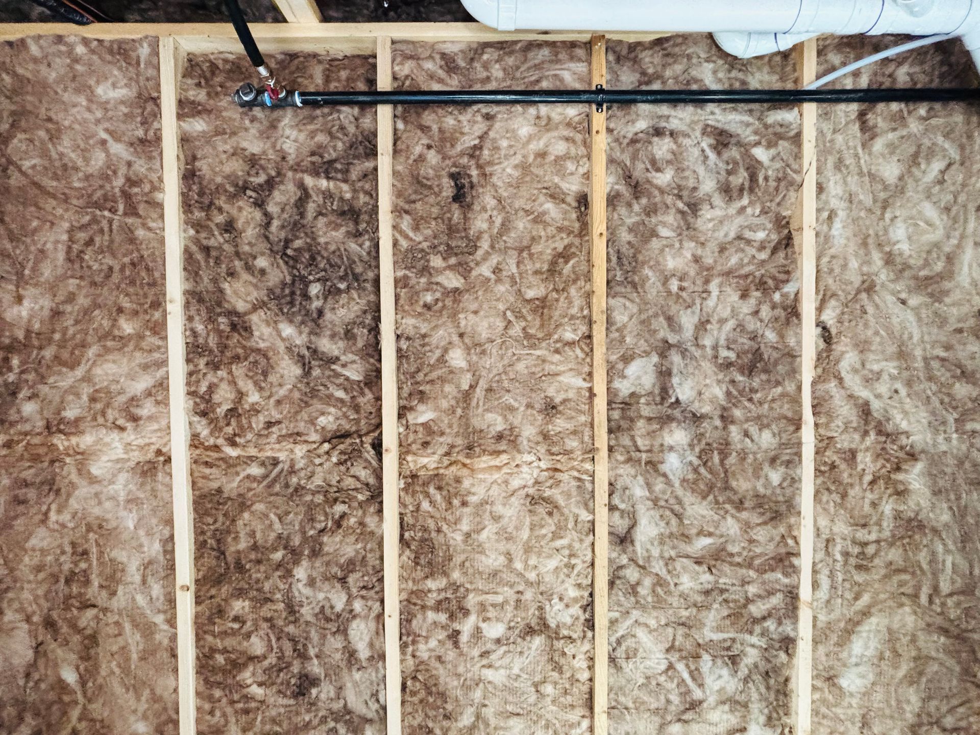 Wall insulation with wooden studs, a black pipe, and sections of dark staining.