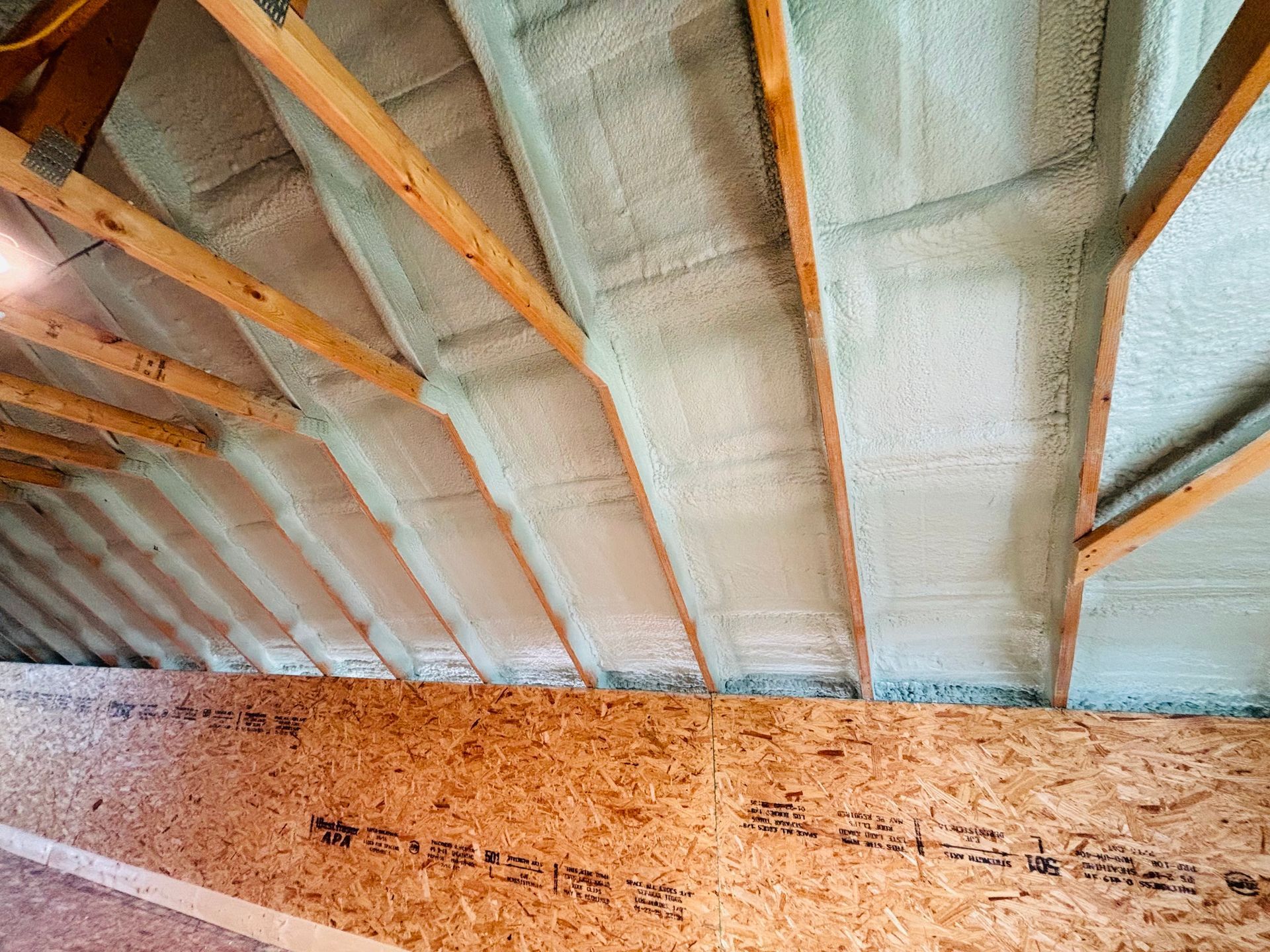 Attic interior with spray foam insulation between wooden rafters and on sheathing; light tan color.