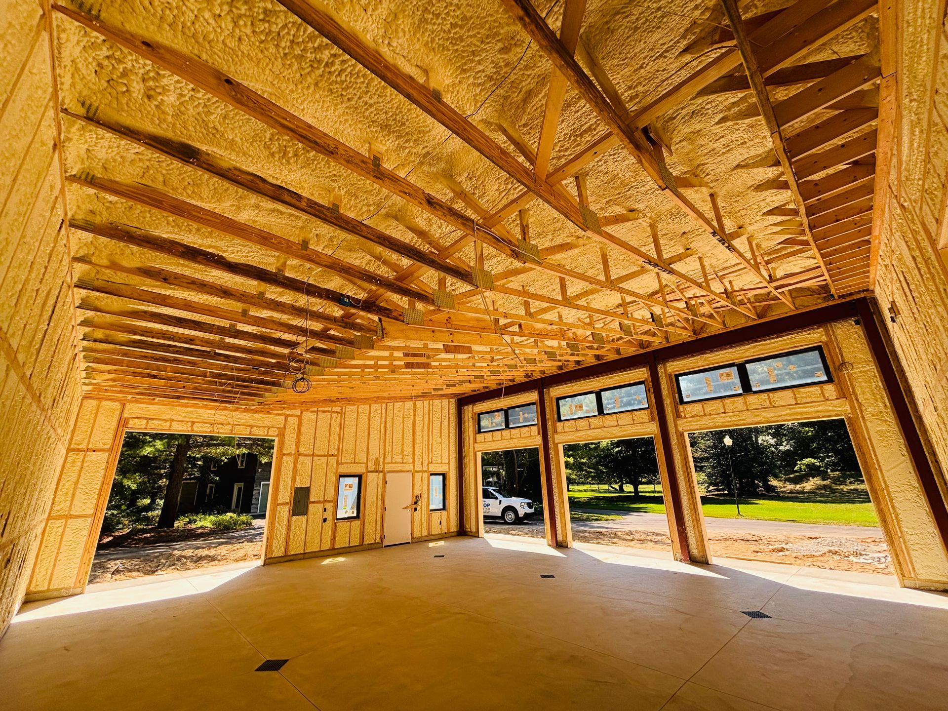 Interior view of a building under construction, spray foam insulation on walls and ceiling. Wooden beams, windows, and concrete floor.