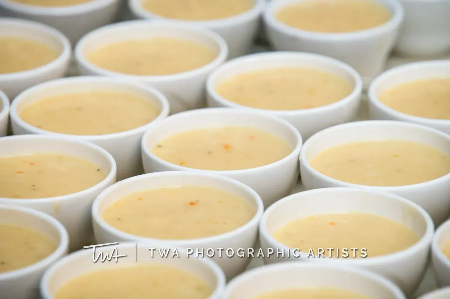 Rows of white cups filled with pale yellow soup or custard on a table