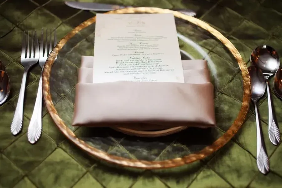 Elegant place setting with gold-rimmed charger, folded napkin, menu card, and silverware on green tablecloth
