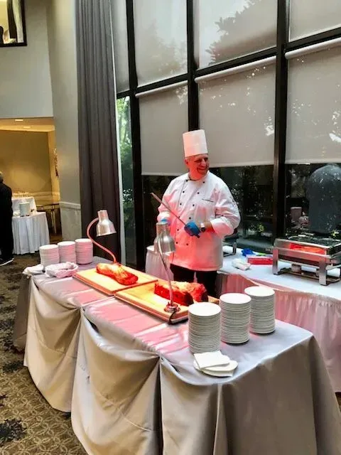 Chef in white uniform serving food at a buffet table with trays and stacked bowls by tall windows