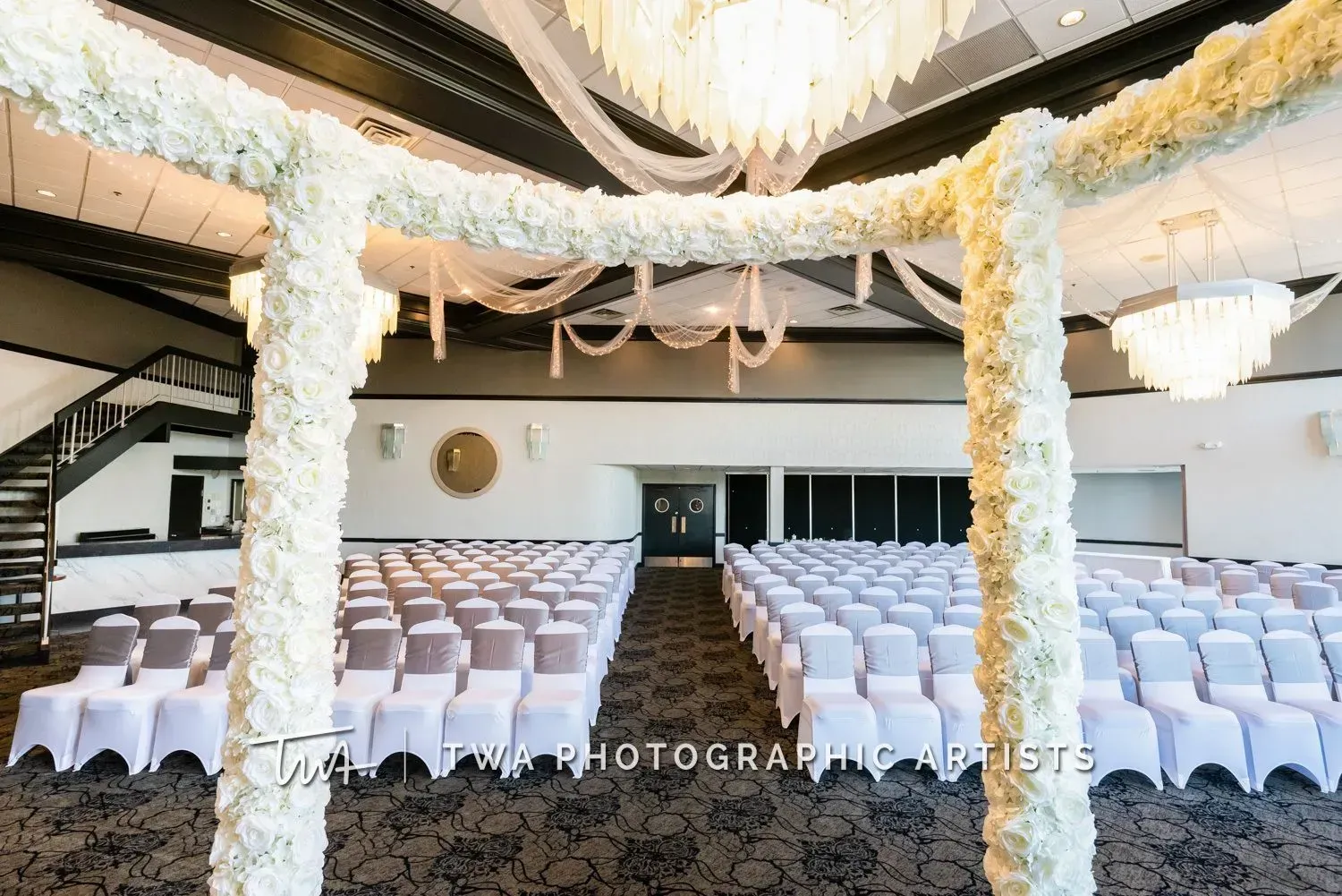 Decorated banquet hall with rows of white-covered chairs under floral drapes and chandeliers