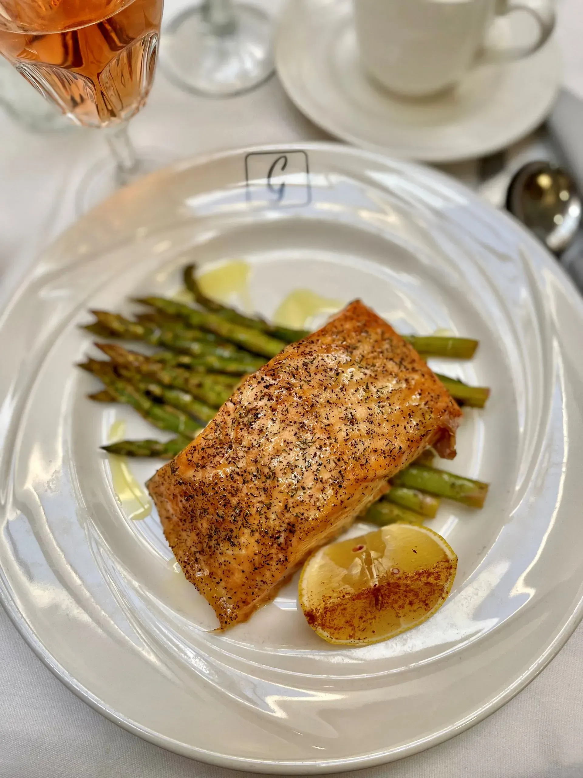 Pan-seared salmon with asparagus and lemon sauce on a white plate, set on a table with tea cups.