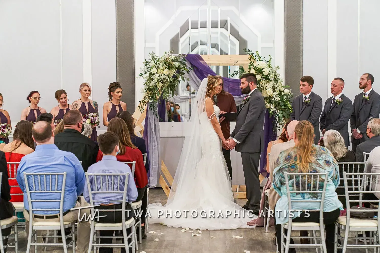 Wedding couple exchanging vows under a purple arch in a bright ceremony hall, with guests seated on both sides.