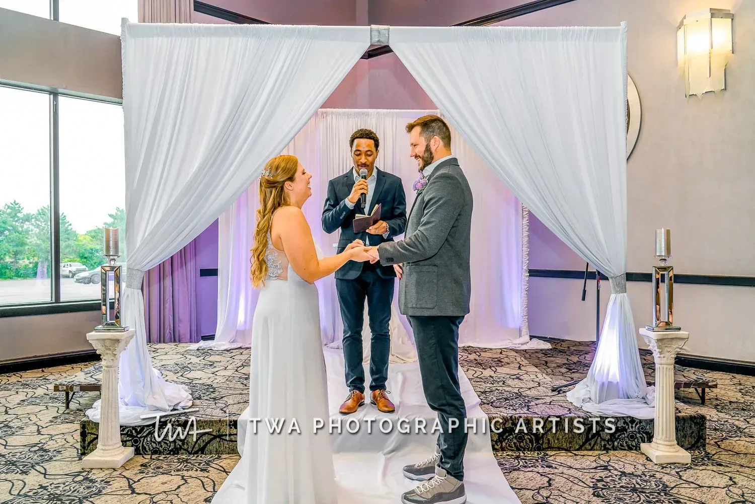 Bride and groom exchange vows under a white draped arch with an officiant at an indoor wedding ceremony.