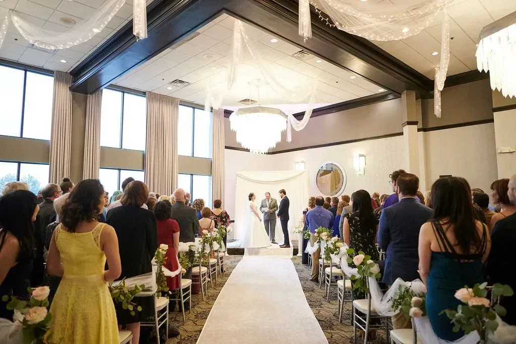 Indoor wedding ceremony with guests seated in a bright hall under draped chandeliers, facing the altar.