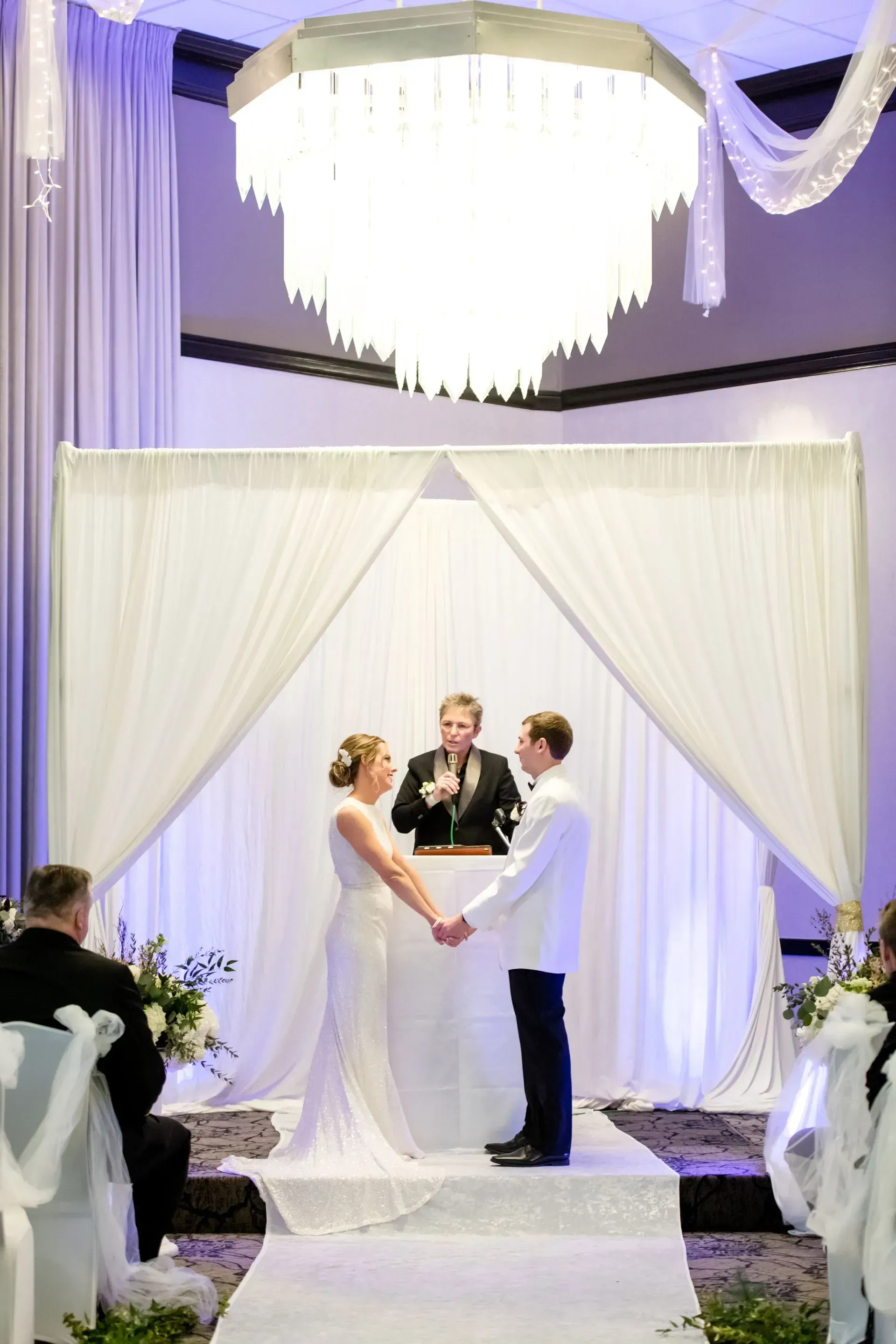 Wedding ceremony under a white draped arch with a couple holding hands as an officiant stands between them.