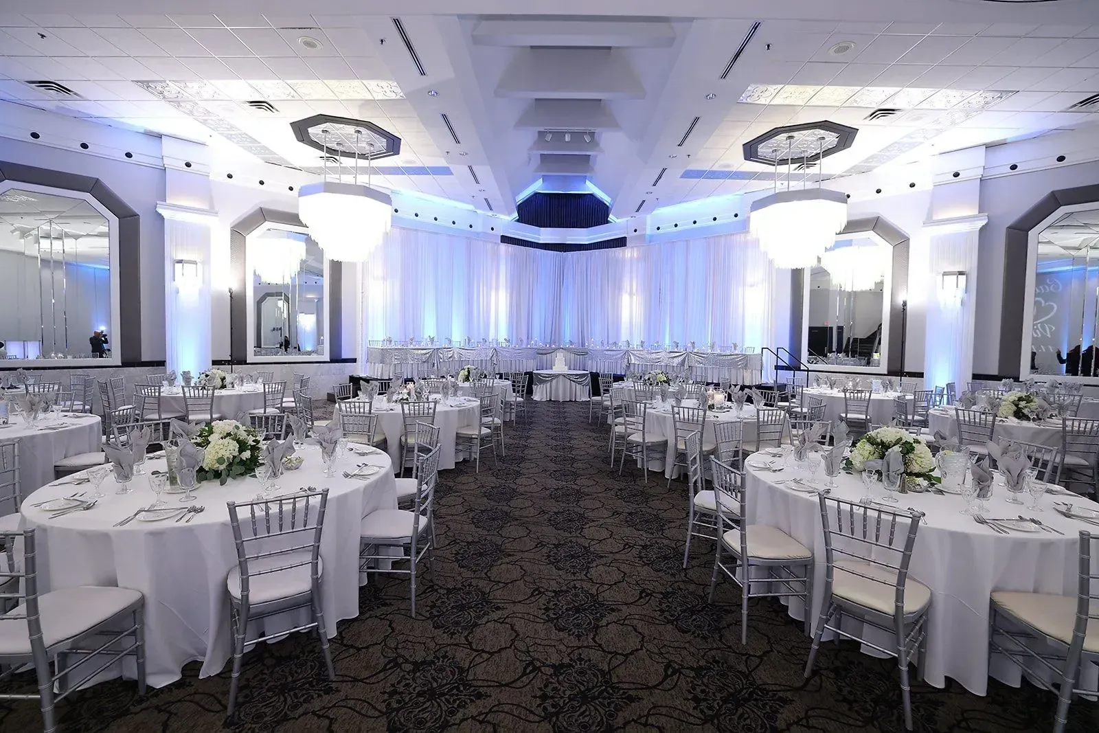 Elegant banquet hall with round white-tablecloth tables, chandelier lighting, and blue uplighting