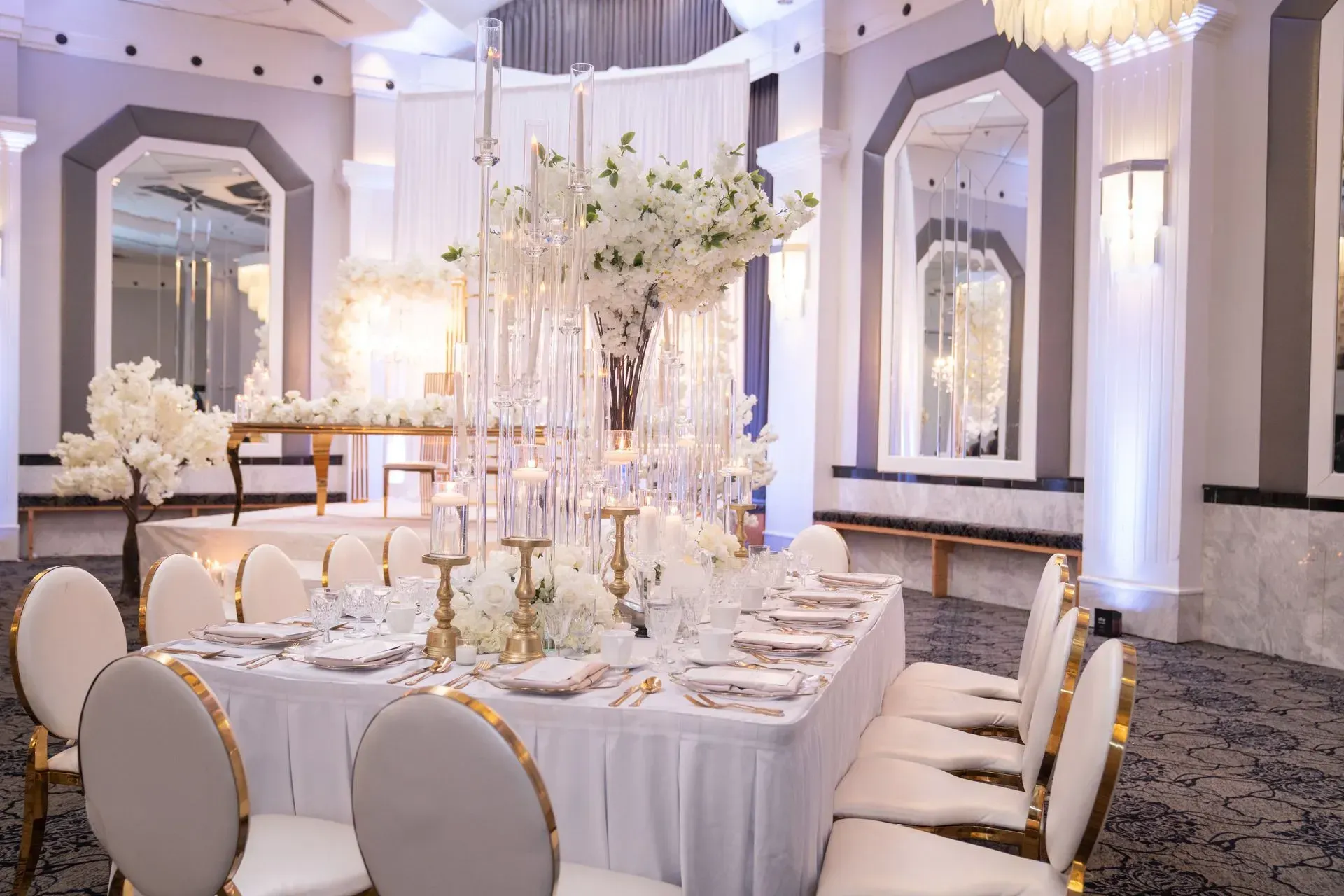 Elegant white banquet hall with round tables, gold chairs, and tall floral centerpieces under soft lights