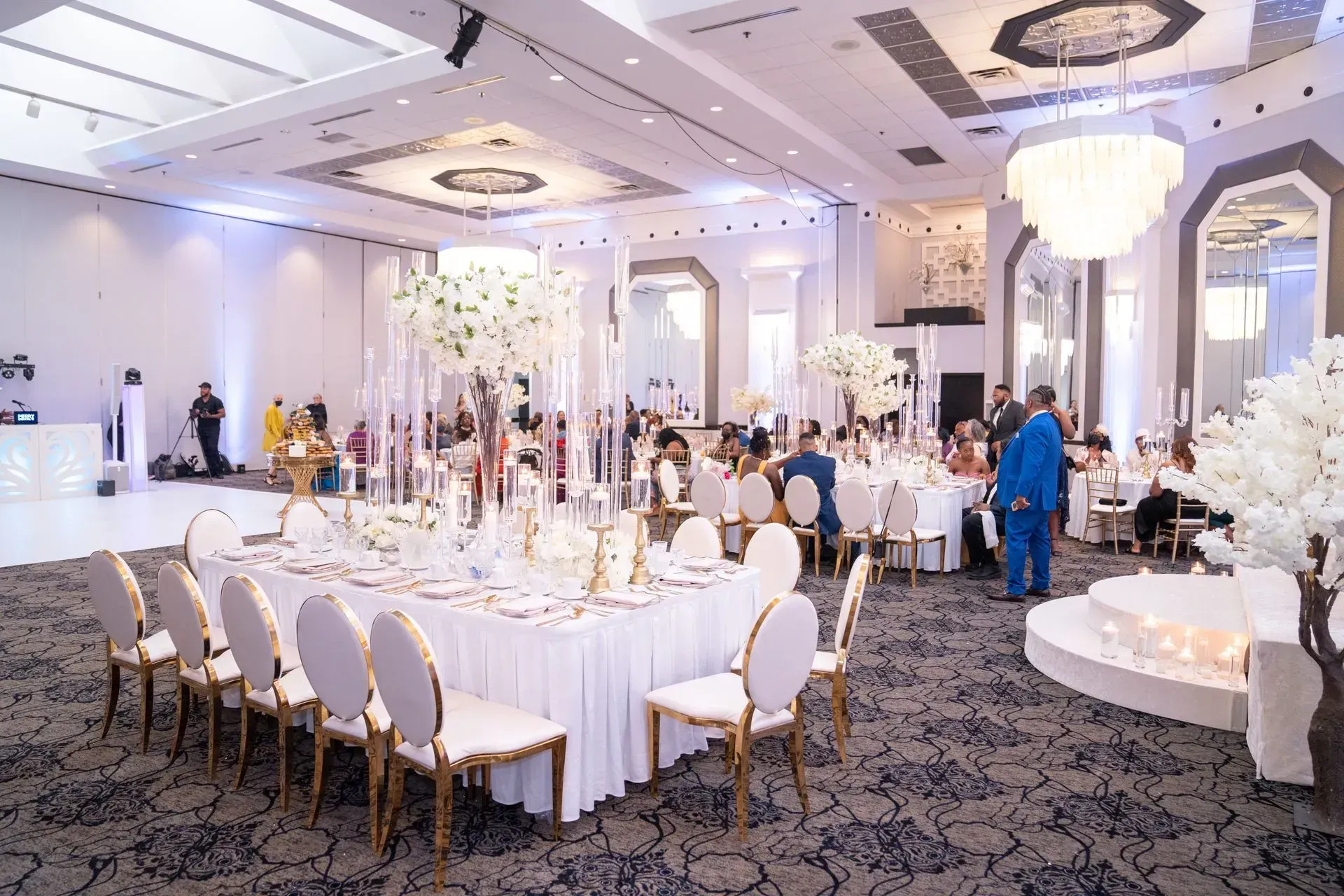 Elegant banquet hall with round tables, white chairs, chandeliers, and guests at a formal event