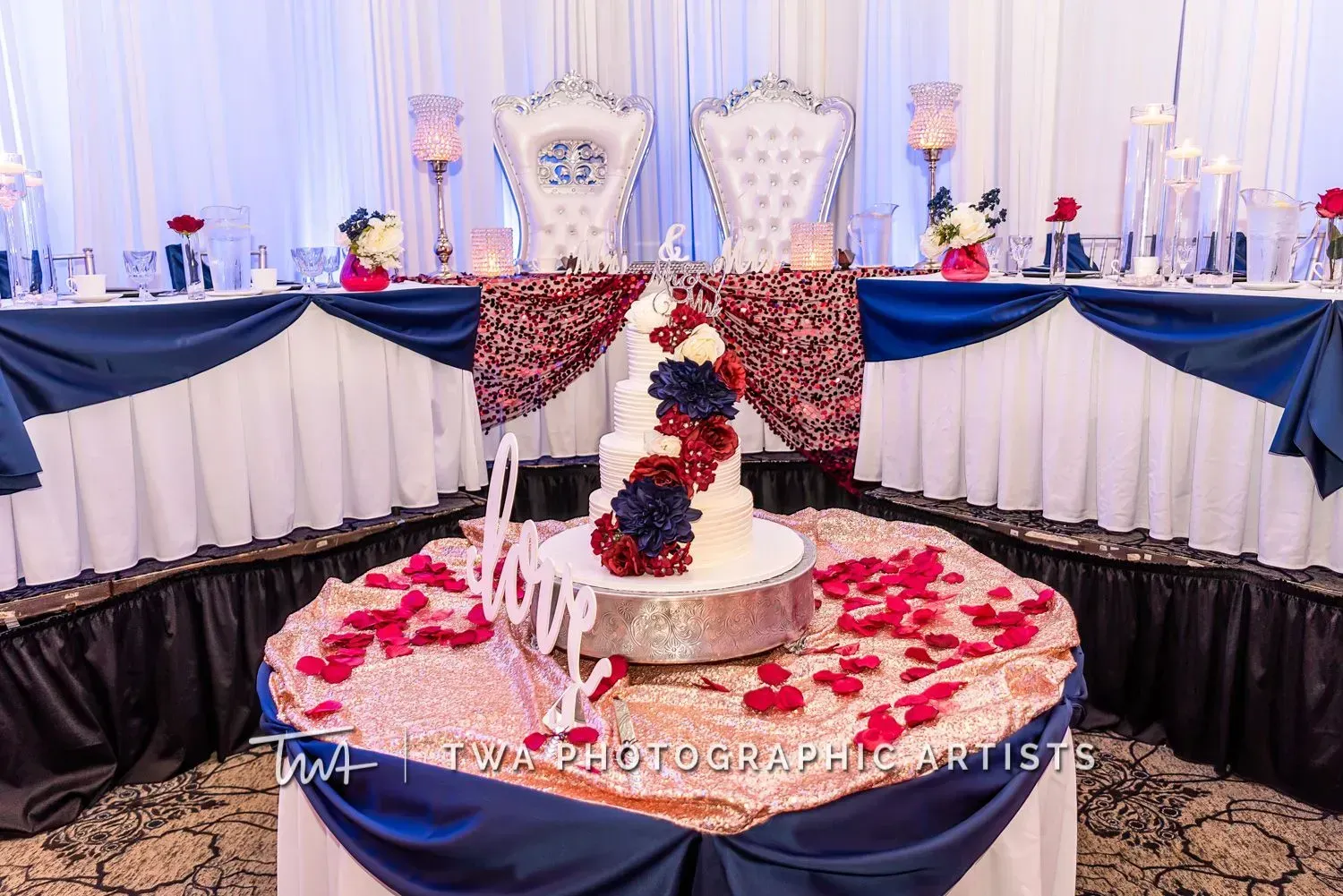 Elegant dessert table with white-and-red cake, rose petals, and gold chairs under blue-and-white drapery