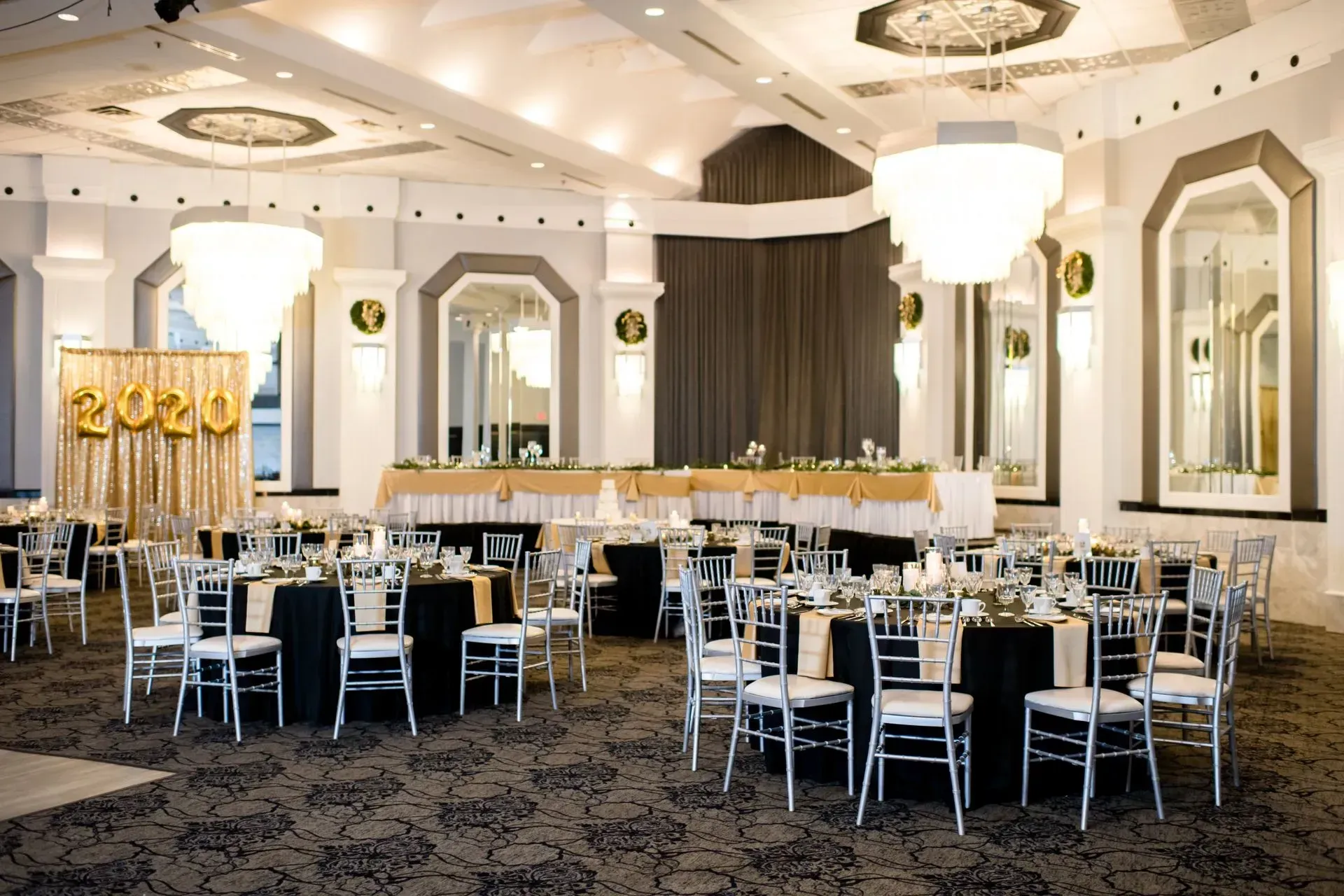 Elegant banquet hall with round tables, white chairs, and gold “2020” decor on the stage
