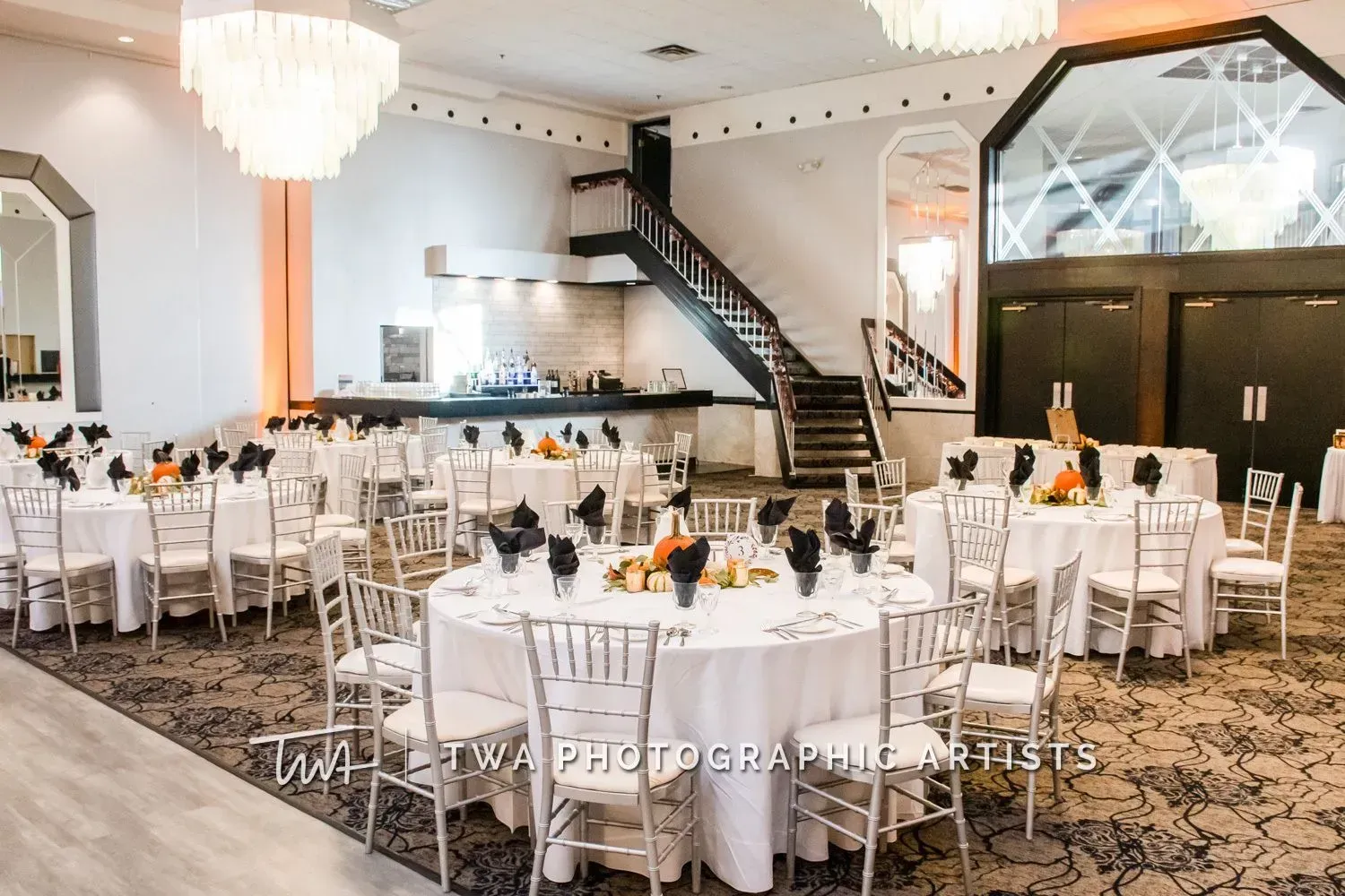 Elegant banquet hall with round white tables, chandeliers, and a staircase at the back.