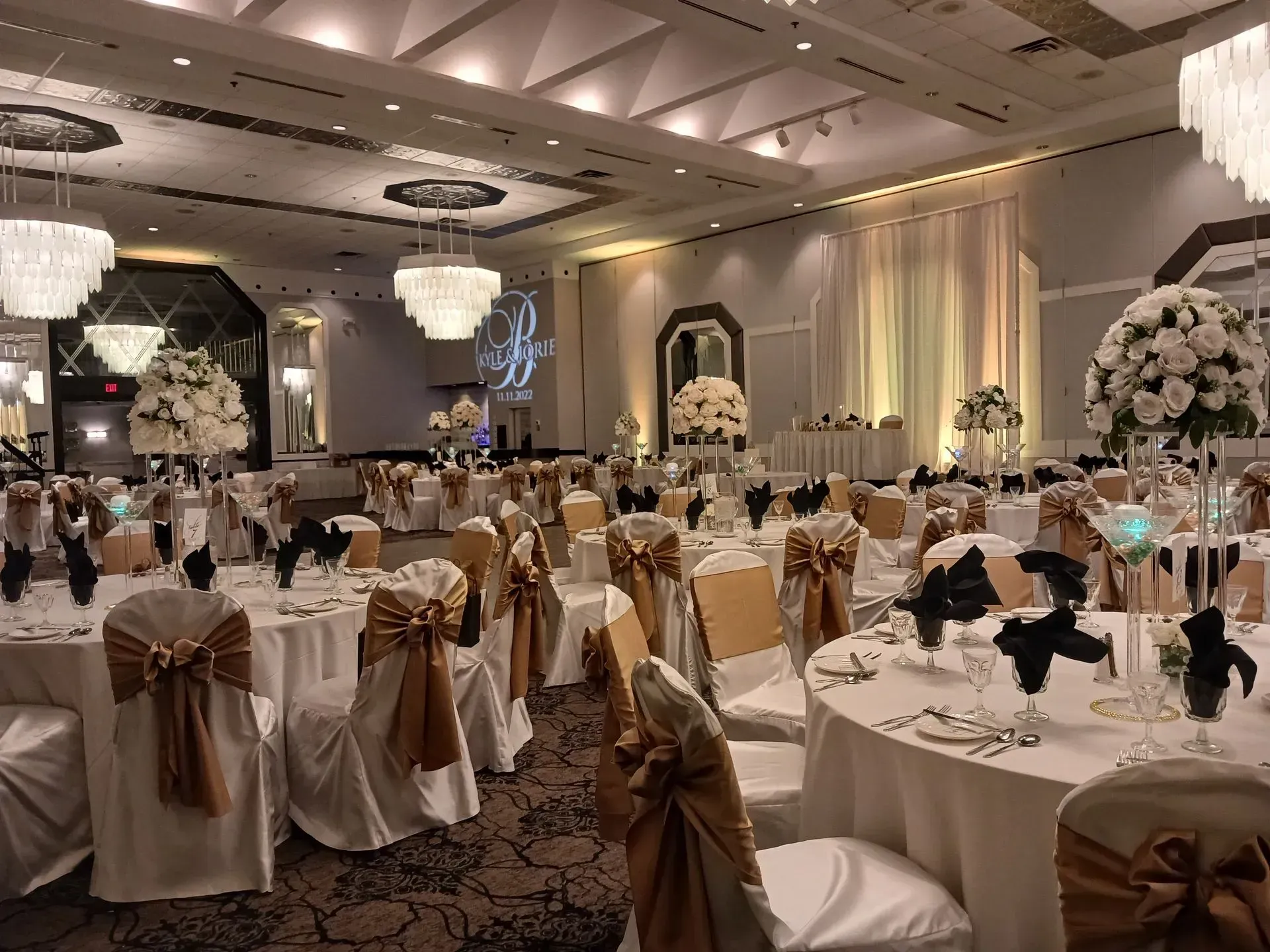Elegant banquet hall with round tables, gold chair bows, white floral centerpieces, and chandeliers.