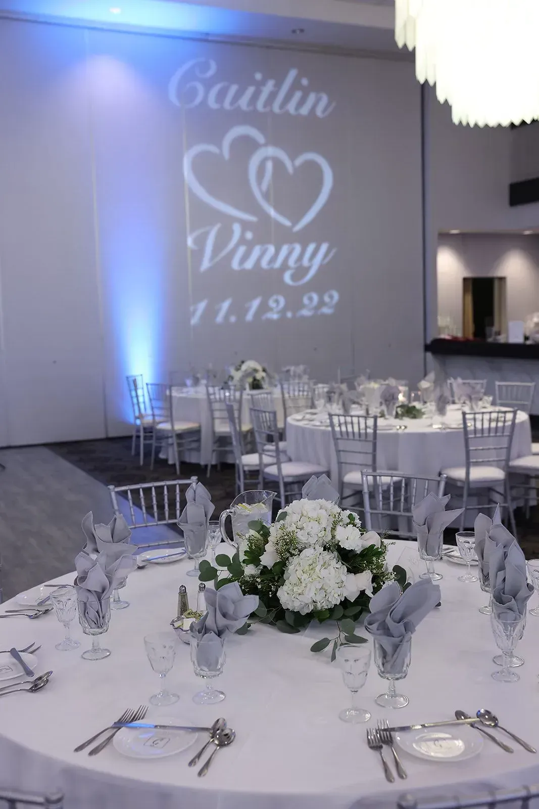 Elegant wedding reception table with white linens, silver decor, and a projected “Caitlin & Vinny 11.19.22” monogram