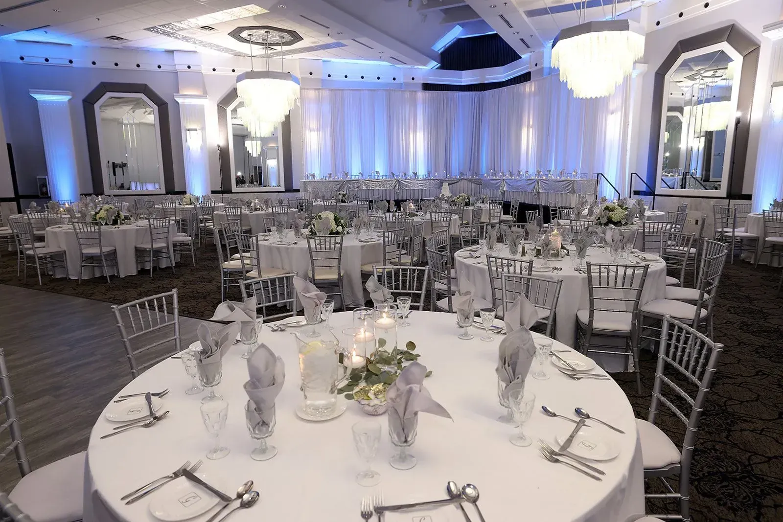 Elegant banquet hall with round, set tables, white decor, and blue uplighting beneath a high ceiling