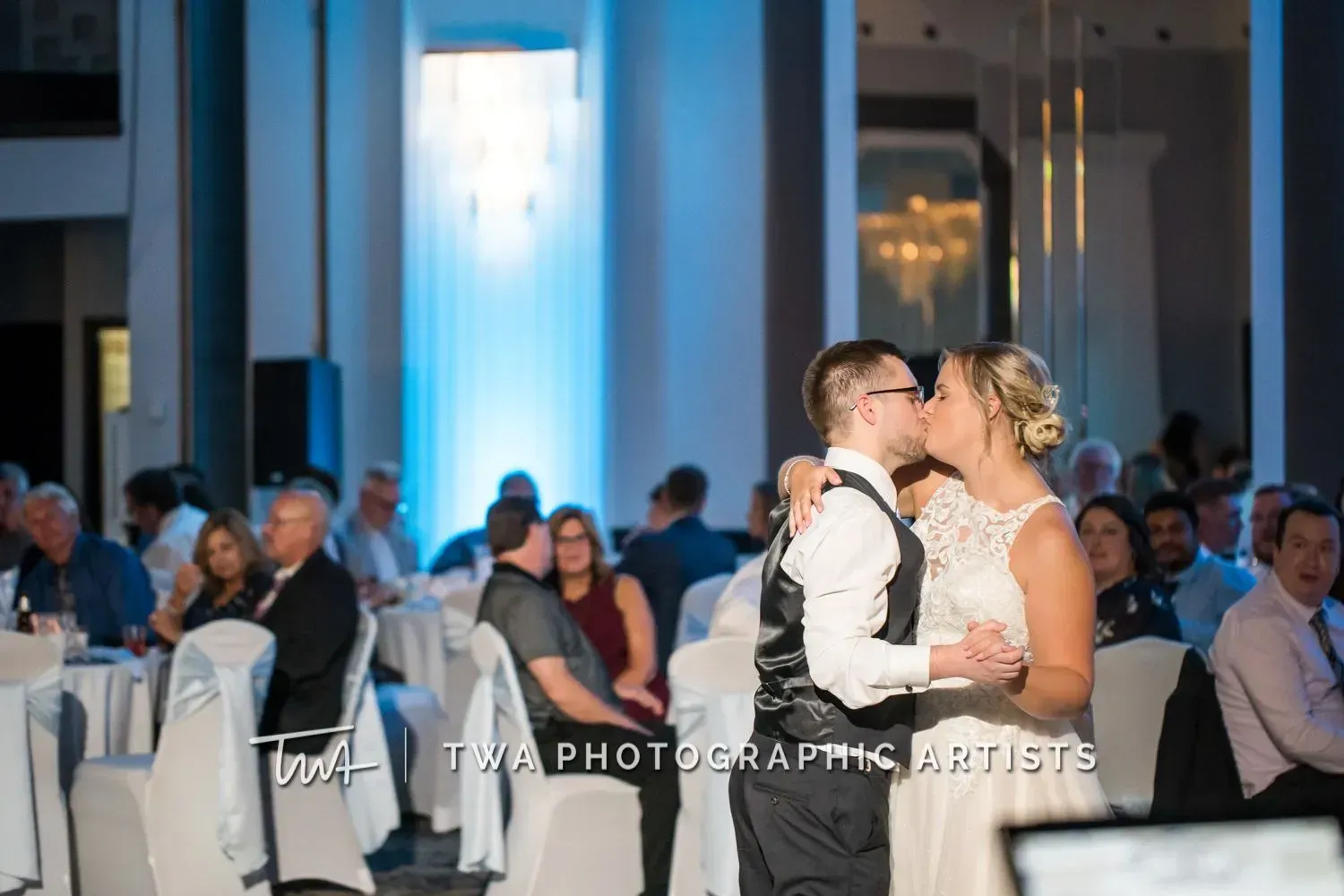 Couple kissing on a dance floor at a formal event, with seated guests and blue lighting in the background