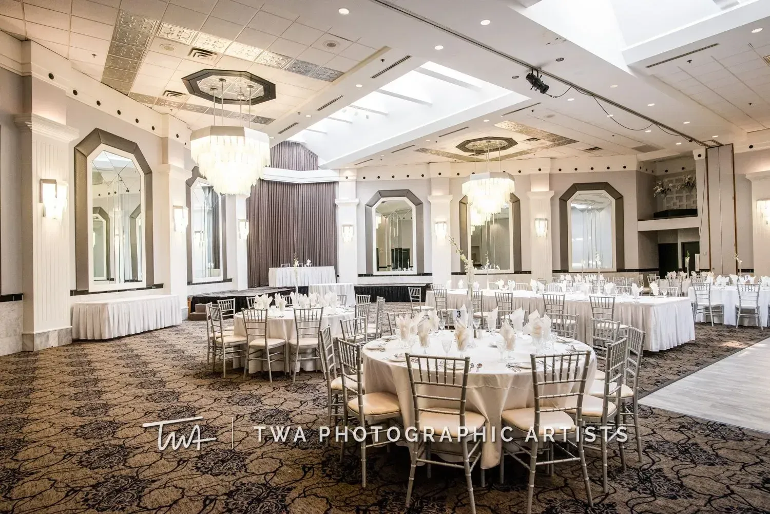 Elegant ballroom with round banquet tables, white chairs, chandeliers, and mirrored wall panels
