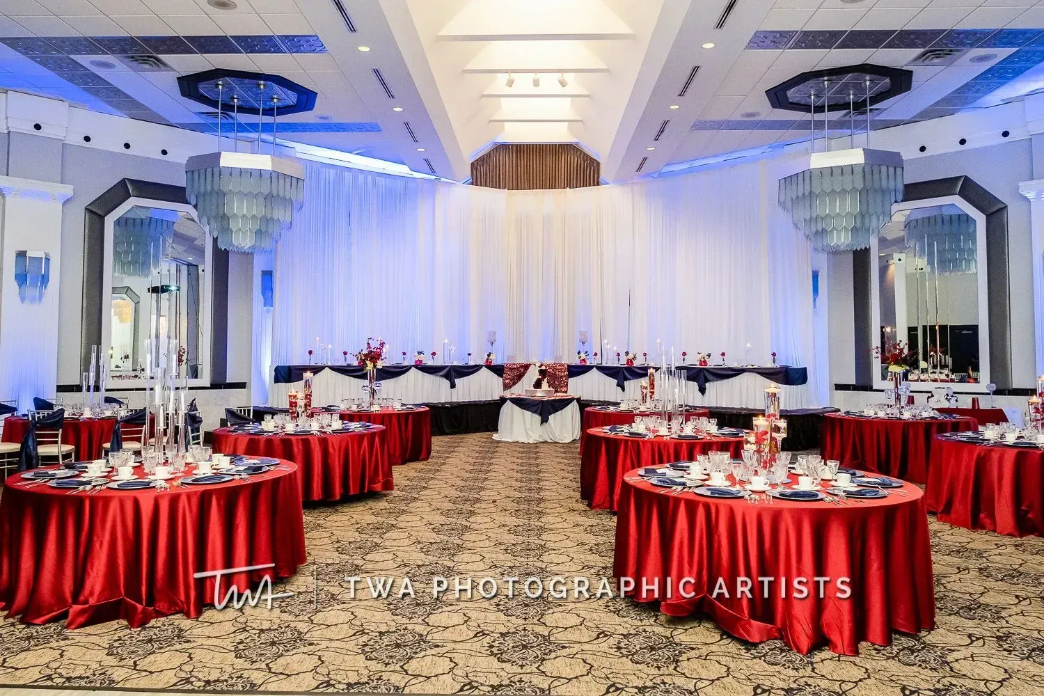 Elegant banquet hall with red round tables, white drapes, and blue ceiling lights