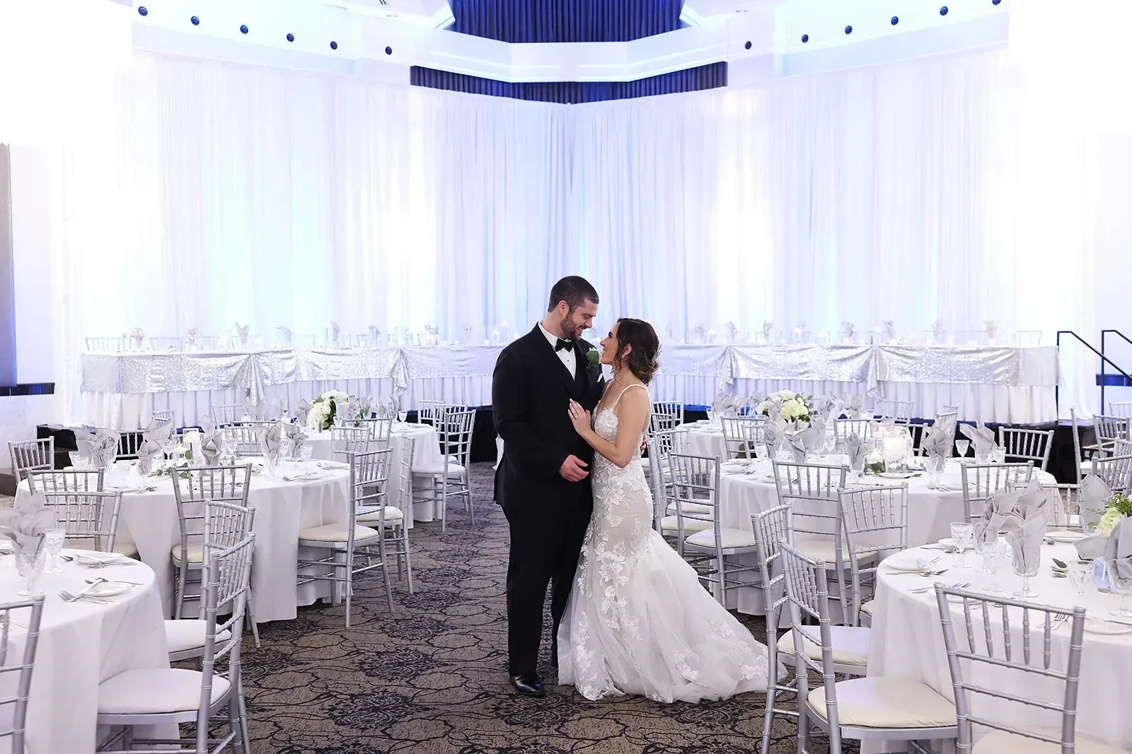 Bride and groom dancing in a white wedding reception hall surrounded by empty tables and chairs