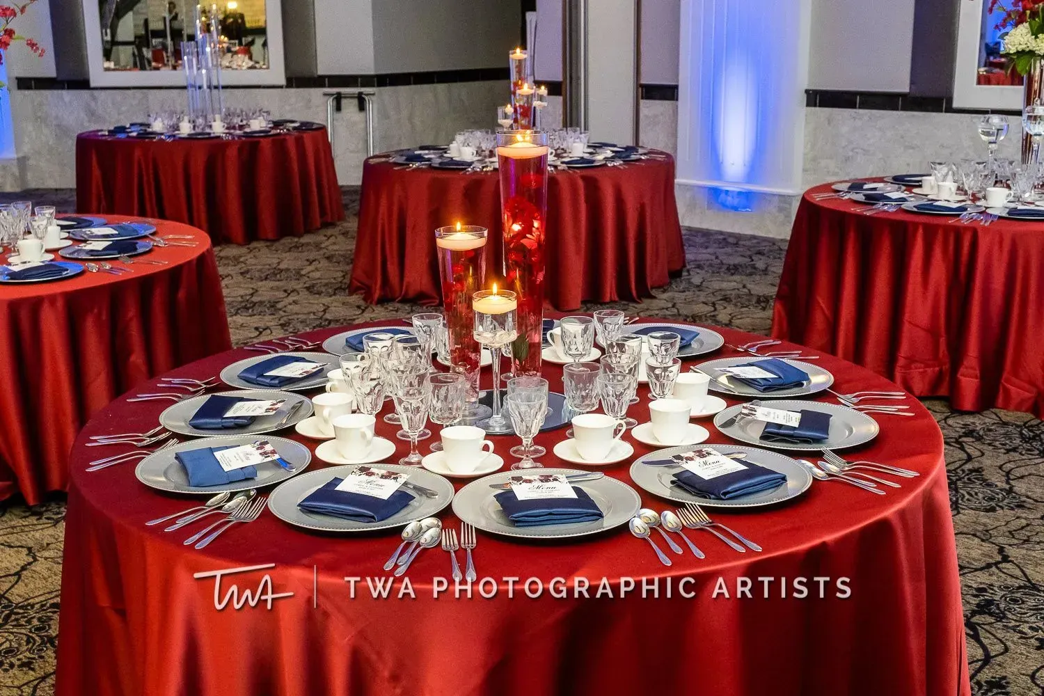Elegant banquet tables with red tablecloths, white plates, and candle centerpieces in a decorated event hall