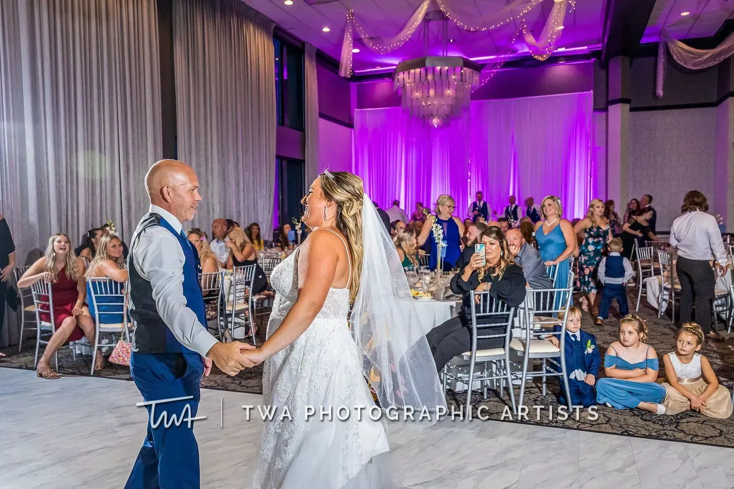 Bride and groom dancing at a wedding reception with purple-lit drapes and seated guests in the background