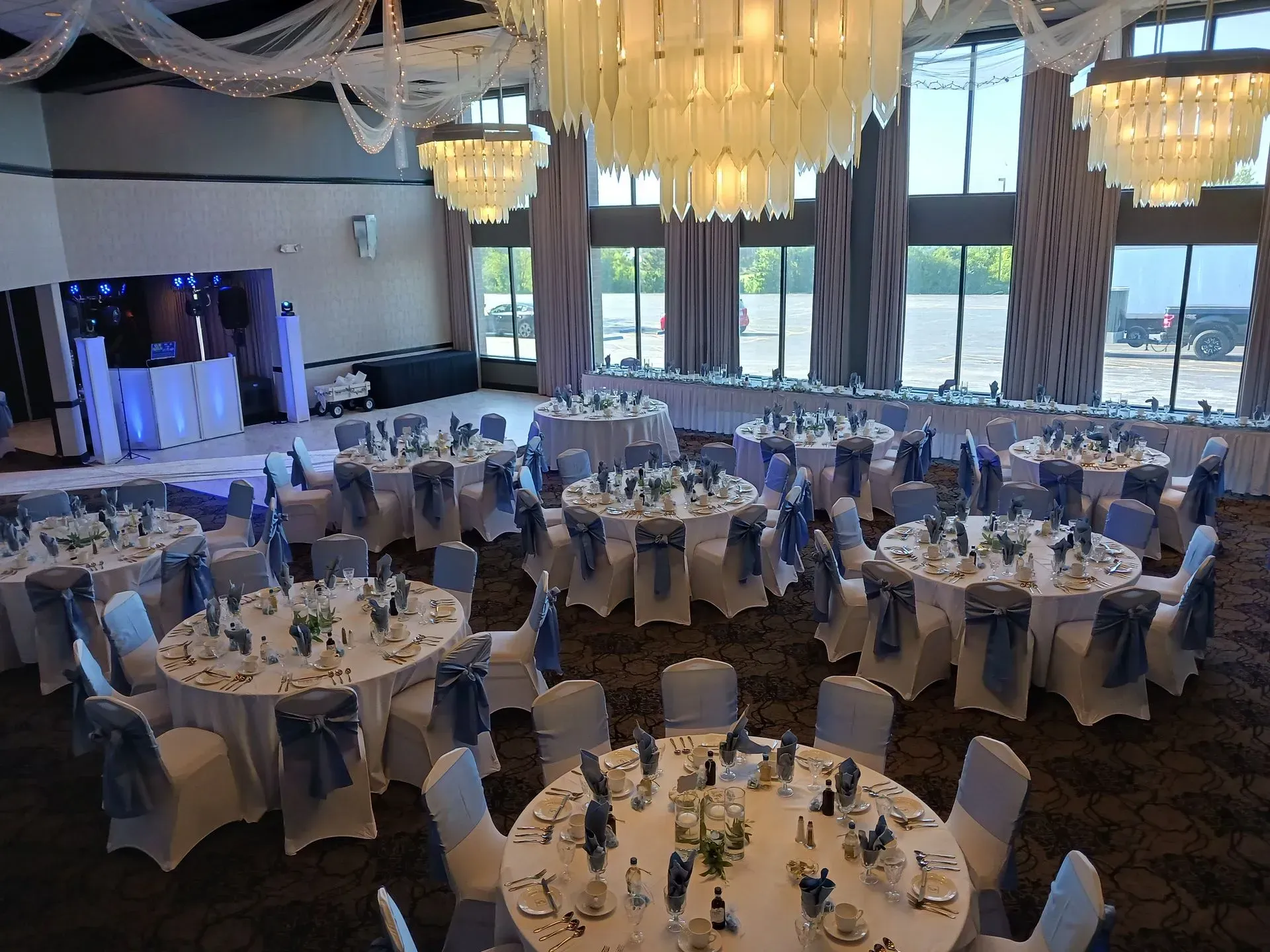 Elegant banquet hall with round tables, white chairs, blue table settings, and large chandeliers by tall windows